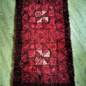 May include: A rectangular, shaggy rug in shades of red and burgundy. The rug features a repeating pattern of squares and stylized white crosses. The edges are fringed, adding texture to the design. This rug would be suitable for a living room or bedroom.