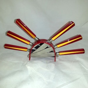 Puede incluir: Un juego de seis cuchillos con mangos rojos y amarillos con un soporte de metal rojo a juego. Los cuchillos están dispuestos en forma de abanico.