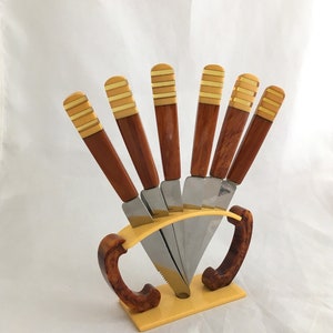 Puede incluir: Juego de siete cuchillos vintage con mangos marrones y dorados en un soporte de plástico amarillo y marrón.