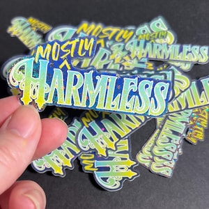 Op de afbeelding: Een stapel holografische stickers met de tekst "Mostly Harmless" in een blauw en geel kleurenschema. De stickers hebben een glinsterende afwerking.