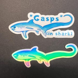 May include: Two shark stickers. The top sticker is blue with the text "* Gasps * (in shark)". The bottom sticker is a rainbow gradient. Both stickers have a white border and are on a black background.