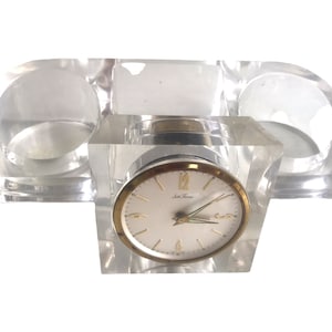 May include: A clear acrylic desk organizer with two cylindrical compartments and a clock in the center. The clock face is white with black Roman numeral hour markers and black hands. The clock face has the text "Jule Time" printed on it.