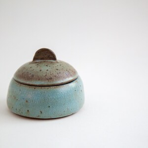 May include: A small, round ceramic lidded jar featuring a speckled glaze. The base is a light blue color with dark brown speckles, while the domed lid transitions from a light blue to a darker brown at the top, finished with a small handle.