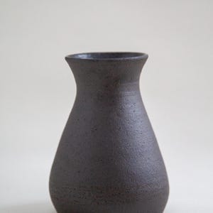 May include: A dark charcoal-colored ceramic vase with a textured surface. The vase has a rounded body and a flared, narrow neck. The handmade pottery has a matte finish and is suitable for home decor.