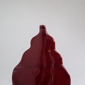 May include: A deep red ceramic vase with a glossy finish and a distinct, organic, multi-tiered silhouette. The vessel features a narrow neck at the top and a wide, rounded base, showcasing a smooth, reflective surface and a flat, unglazed bottom edge.