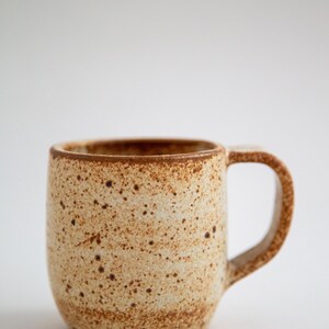 May include: A ceramic mug with a speckled tan and brown finish. The mug features a cylindrical body with a rounded base and a sturdy, curved handle on the right side. The surface has a textured appearance with dark brown spots throughout the clay.