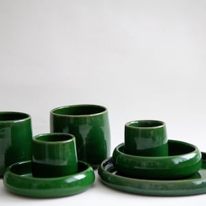 Könnte beinhalten: Eine Sammlung glänzender, smaragdgrüner Keramikgeschirr. Das Set umfasst zylindrische Tassen, kleine Schalen und Teller. Die Teile haben eine glatte, reflektierende Oberfläche und sind auf weißem Hintergrund angeordnet. Das Geschirr eignet sich zum Servieren von Speisen.