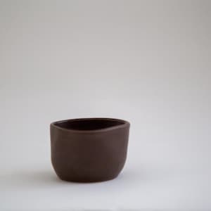 May include: A small, dark brown ceramic cup with a rounded shape and a slightly irregular rim. The cup is set against a plain white background, highlighting its simple, minimalist design. The cup is approximately 2 inches in diameter.