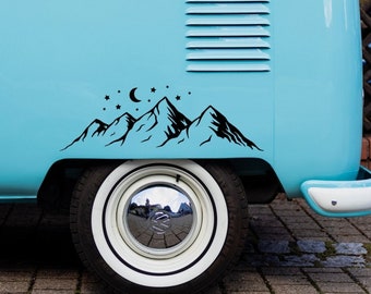 May include: A blue and white vintage van with a black mountain range and stars decal on the side. The van has a white roof rack and a white tire with a chrome hubcap.