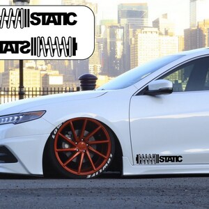 May include: A white car with a black "STATIC" decal on the side. The decal features a stylised image of a coil spring. The car has orange wheels and is parked on a street.