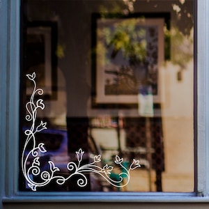 May include: White floral design sticker on a glass window. The design features a vine with leaves and flowers, creating a corner border.