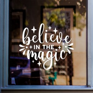 May include: White vinyl decal with the text "Believe in the magic" with stars and swirls around the text. The decal is on a glass window.