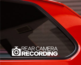 May include: White vinyl sticker with a camera icon and the text "REAR CAMERA RECORDING" on a red car window.