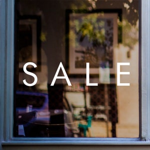 May include: A shop window with the word "SALE" displayed in white lettering on the glass. The window reflects the interior of the shop, which is blurred.