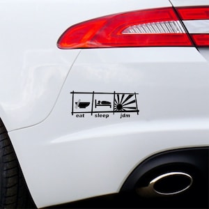 May include: Black and white sticker on a white car bumper. The sticker shows a stylized illustration of a bowl of noodles, a bed, and a rising sun with the words "eat", "sleep", and "jdm" underneath.