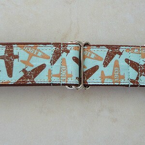 May include: A dog collar with a light blue background and a repeating pattern of brown and tan airplanes. The collar has a brown border and silver hardware.