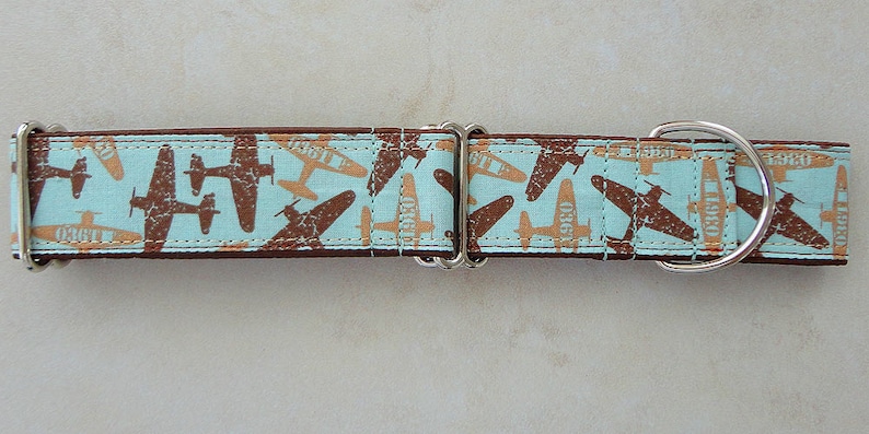 May include: A dog collar with a light blue background and a repeating pattern of brown and tan airplanes. The collar has a brown border and silver hardware.