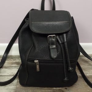 May include: Black Esprit backpack with a flap closure and a small top handle. The backpack has adjustable straps and a front zipped pocket. The Esprit logo is embossed on the flap. The bag is made of a textured material.