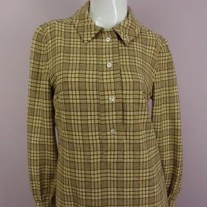 May include: A long-sleeved shirt featuring a tan and brown plaid pattern. The garment has a pointed collar, a button-down placket with four visible white buttons, and cuffed sleeves. The fabric displays a consistent grid-like check design.