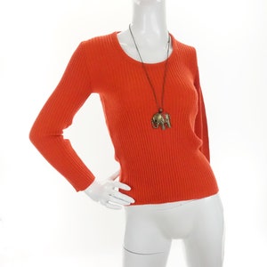 May include: A bright orange ribbed jumper with long sleeves and a round neckline. The jumper is worn by a mannequin and features a gold elephant pendant necklace.