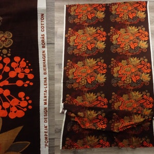 May include: A brown fabric with a repeating pattern of orange and brown flowers. The fabric has a white label with the text "POMPEJA DESIGN MARTA-LENA BJERHAGEN BOR&Aring;S COTTON".