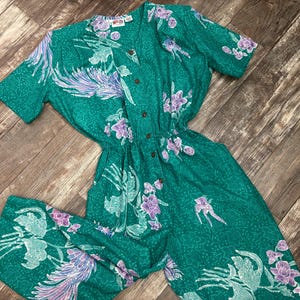 May include: A teal jumpsuit with a floral pattern in shades of purple and white. The short-sleeved jumpsuit features a button-down front and an elastic waist. The fabric has a subtle textured pattern.