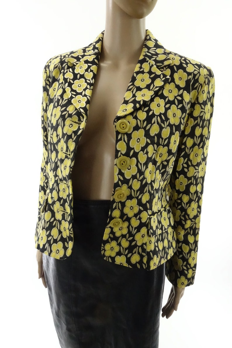Puede incluir: Una chaqueta negra con estampado floral amarillo con cierre de botones. La chaqueta tiene una silueta ajustada y mangas largas.