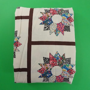 May include: White fabric with a brown border and a repeating pattern of colourful patchwork stars. The stars are made of different coloured fabrics and have a central circle.