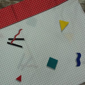 May include: A white fabric with a grid pattern, featuring colourful geometric shapes: a blue square, a green square, a yellow triangle, a red triangle, and a blue wavy shape. A red and white checkered border is at the top.