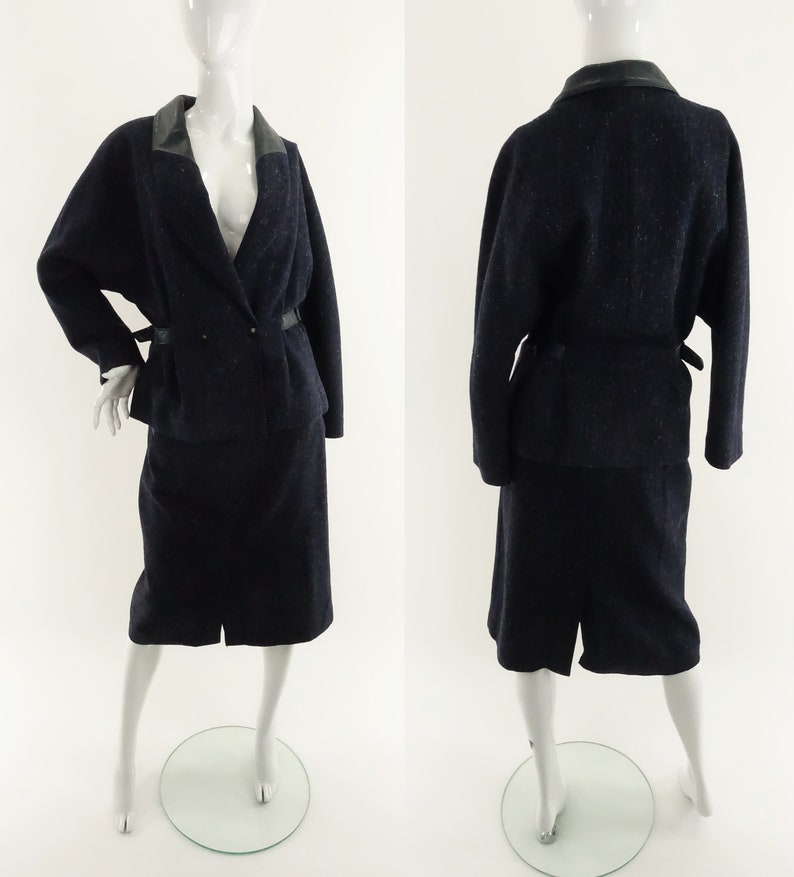 May include: A navy blue skirt suit with a black leather collar and a matching belt. The suit is made of a textured fabric and has a fitted silhouette.