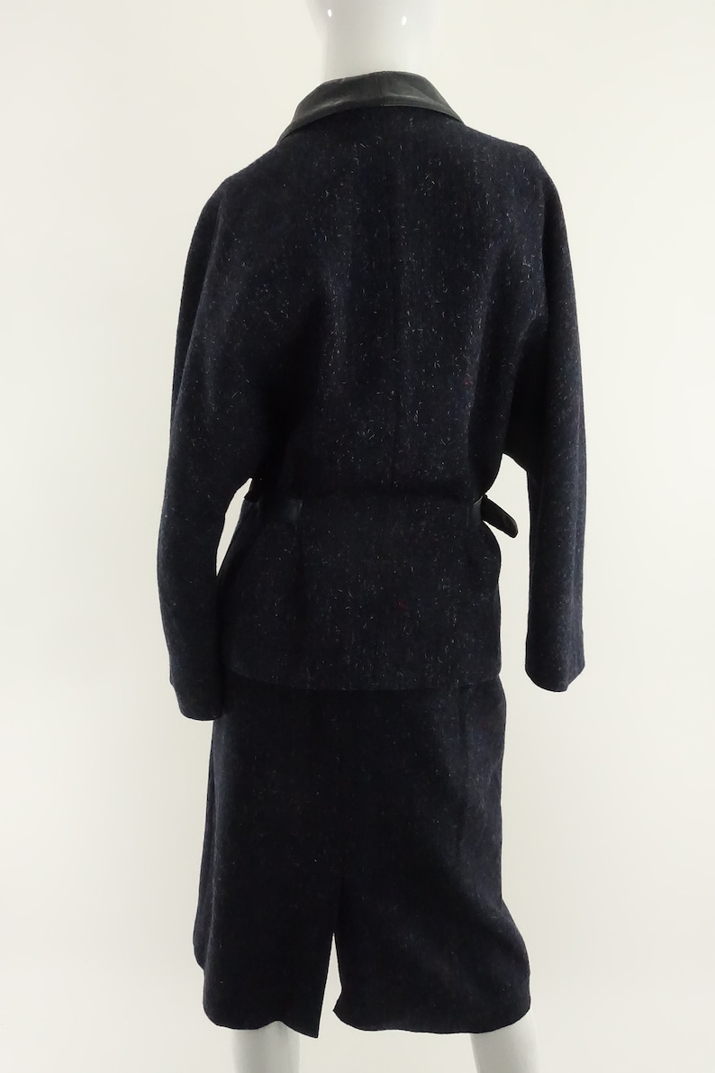 May include: A navy blue wool suit with a black leather collar and a matching belt. The jacket has long sleeves and a relaxed fit. The skirt is knee-length and has a straight silhouette.