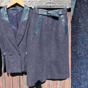 May include: A blue tweed skirt suit with a double-breasted jacket and a matching skirt. The jacket has a leather collar and trim. The skirt has a leather belt and trim.