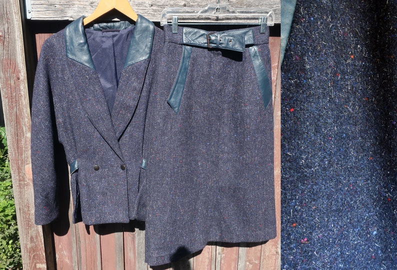 May include: A blue tweed skirt suit with a double-breasted jacket and a matching skirt. The jacket has a leather collar and trim. The skirt has a leather belt and trim.