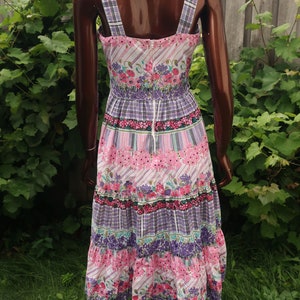 Op de afbeelding: Een roze en paarse bloemenprint midi-jurk met een witte achtergrond. De jurk heeft een getailleerd bovenstuk en een gelaagde rok. De bandjes zijn dun en hebben een ruitpatroon.