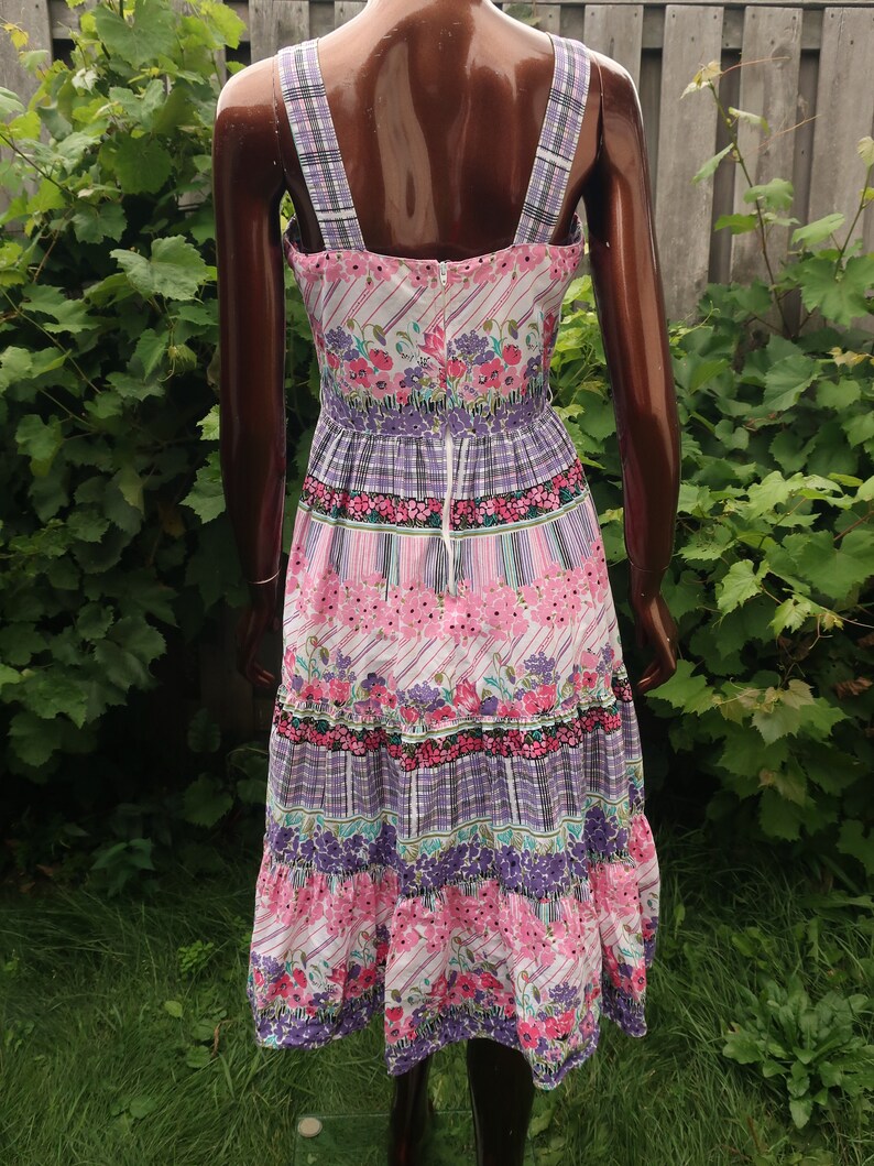 Op de afbeelding: Een roze en paarse bloemenprint midi-jurk met een witte achtergrond. De jurk heeft een getailleerd bovenstuk en een gelaagde rok. De bandjes zijn dun en hebben een ruitpatroon.