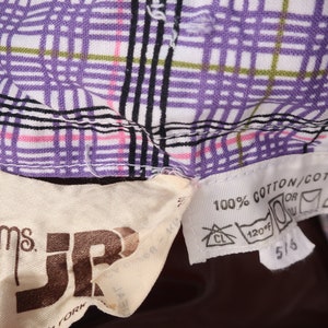 Op de afbeelding: Een close-up van een witte en paarse geruite stof met een bruin label met de tekst "Ms. JR" en een wit label met de tekst "100% Katoen/Coton" met wasinstructies.