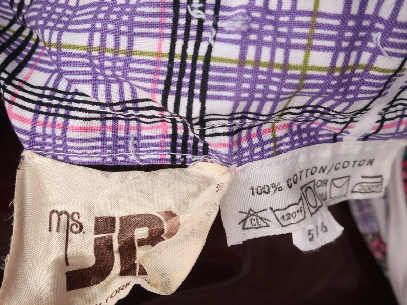 Op de afbeelding: Een close-up van een witte en paarse geruite stof met een bruin label met de tekst "Ms. JR" en een wit label met de tekst "100% Katoen/Coton" met wasinstructies.