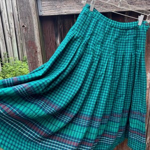 May include: A pleated midi skirt featuring a vibrant green and black tartan pattern with subtle red and white accent lines. The skirt has a high waistband with a single side button closure and is displayed on a clear plastic hanger.