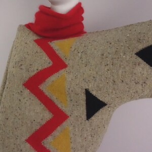 May include: A beige poncho with a red zigzag and yellow triangle design. The poncho has black triangle accents and a red turtleneck collar. The fabric has a speckled texture.