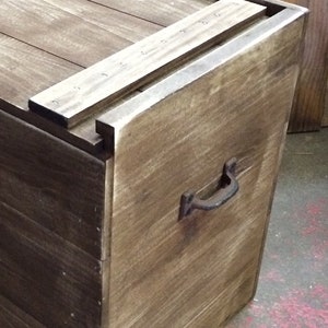 Puede incluir: Una caja de almacenamiento de madera con una tapa con bisagras y una manija de metal. La caja está hecha de madera marrón oscura y tiene un aspecto rústico.