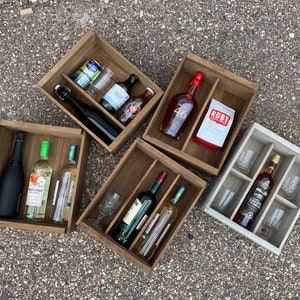 May include: Five wooden crates with dividers, each containing various bottles of alcohol and glasses. The crates are arranged in a semi-circle on a gray asphalt surface.