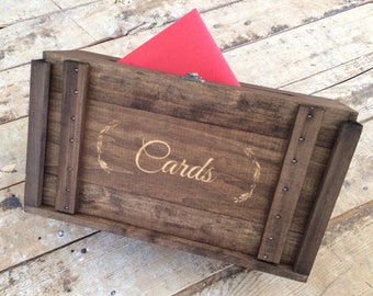 Peut inclure: Une boîte en bois rustique avec un couvercle à charnières, décorée d'une inscription dorée qui dit "Cards". La boîte est posée sur une surface en bois et une enveloppe rouge est partiellement visible à l'intérieur.