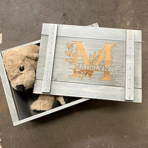 May include: A gray wooden box with a hinged lid. The lid is open and a stuffed animal is inside. The lid has a floral design and the name "Madelyn" engraved on it.