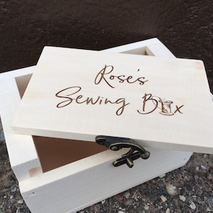 May include: A wooden box with a hinged lid. The lid is open and the box is labeled "Rose's Sewing Box" with a small illustration of a spool of thread and needle.