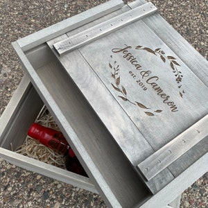 May include: A gray wooden box with a hinged lid. The lid is open and the inside of the box is lined with shredded paper. A red bottle is inside the box. The lid is engraved with the text "Jessica & Cameron est. 2019" and a floral design.