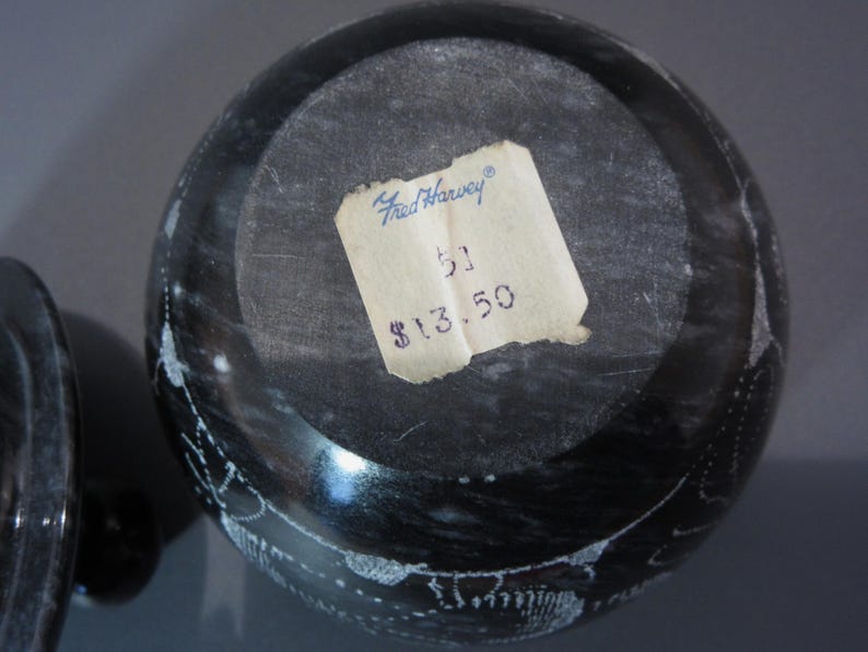 Pu&ograve; includere: Una ciotola rotonda in pietra grigio scuro con coperchio, con venature bianche e un'etichetta di carta. L'etichetta recita "Fred Harvey" e include un numero. La superficie della ciotola &egrave; lucida e riflette la luce.
