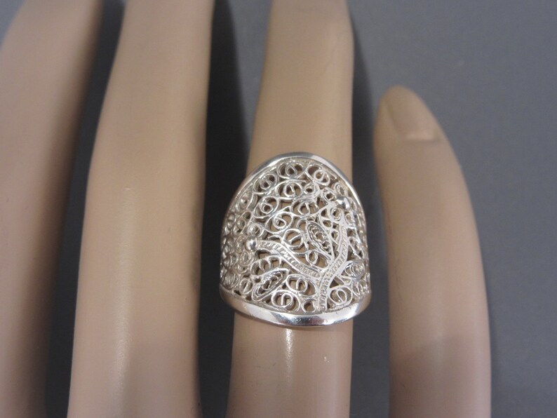 May include: A silver filigree ring with a delicate, intricate design. The ring is wide and has a rounded top.