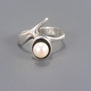 Puede incluir: Un anillo de plata con un dise&ntilde;o abstracto &uacute;nico que presenta una sola perla engastada en un bisel circular.