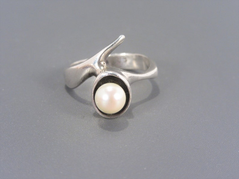 Puede incluir: Un anillo de plata con un dise&ntilde;o abstracto &uacute;nico que presenta una sola perla engastada en un bisel circular.