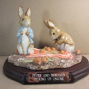 May include: A ceramic figurine set featuring two brown rabbits on a textured base with a red and white checkered cloth and onions. One rabbit stands in a blue jacket, while the other bends over. A plaque reads: PETER AND BENJAMIN PICKING UP ONIONS.